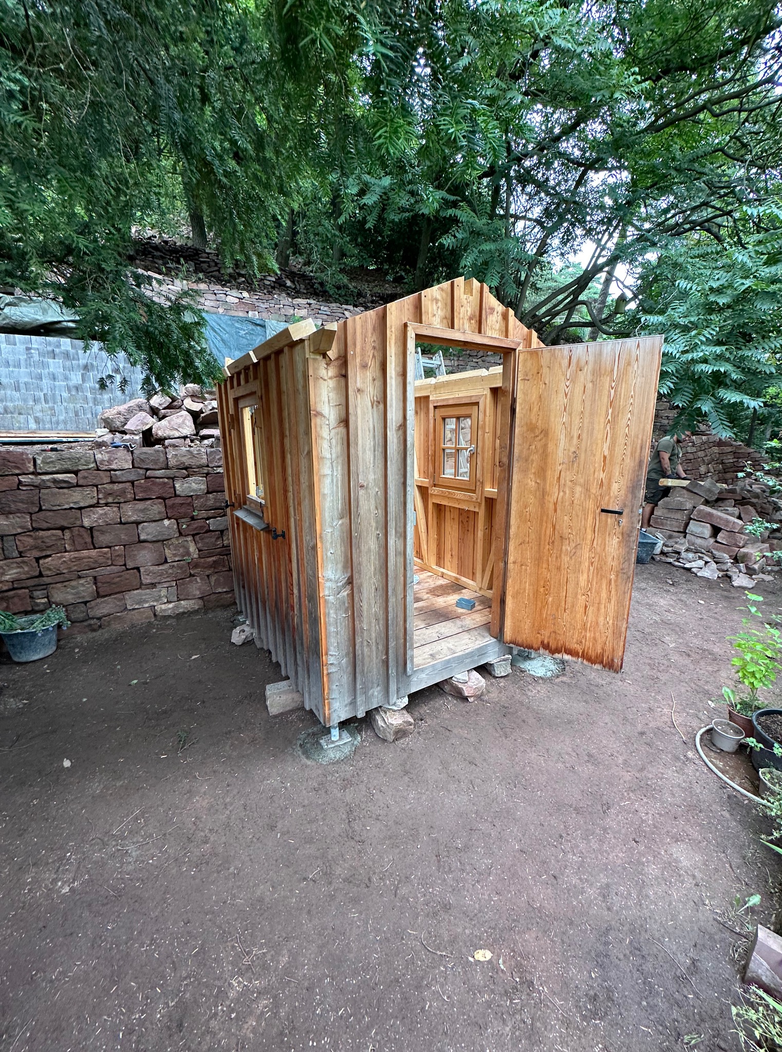 Drewniana altana z otwartymi drzwiami w ogrodzie, otoczona kamiennym murem i zielenią. Widoczne detale konstrukcyjne i naturalna faktura drewna.