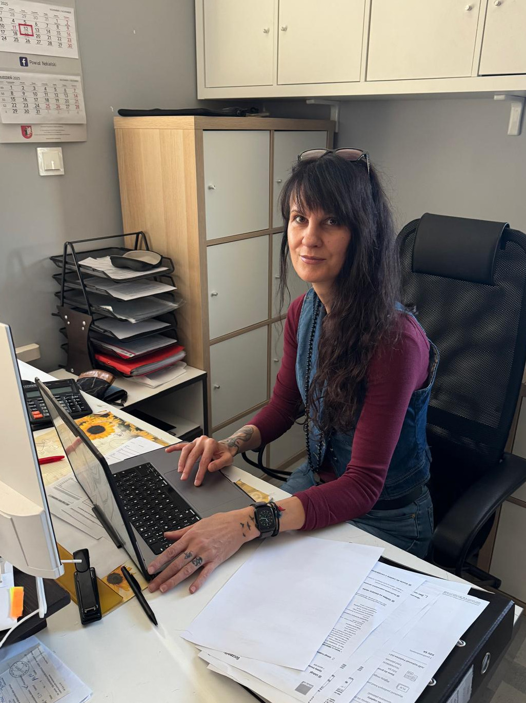 Kobieta z tatuażami przy biurku, pracuje na laptopie. Na biurku dokumenty, kalkulator i przybornik. W tle szafki biurowe i kalendarz. Jasne, naturalne światło.