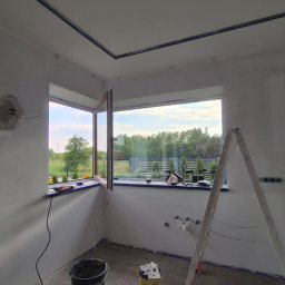 Wimar Usługi Remontowo Budowlane Marcin Kowalonek - Pokój w trakcie malowania: przygotowane ściany, okna z widokiem na zieleń, drabina malarska, narzędzia. Widoczne niedoskonałości na ścianach przed malowaniem.