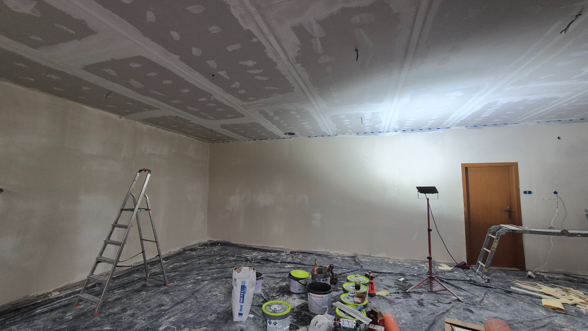 Pokój w trakcie prac wykończeniowych: szpachlowanie sufitu, ściany przygotowane do malowania, widoczne drabiny, farby i narzędzia malarskie.