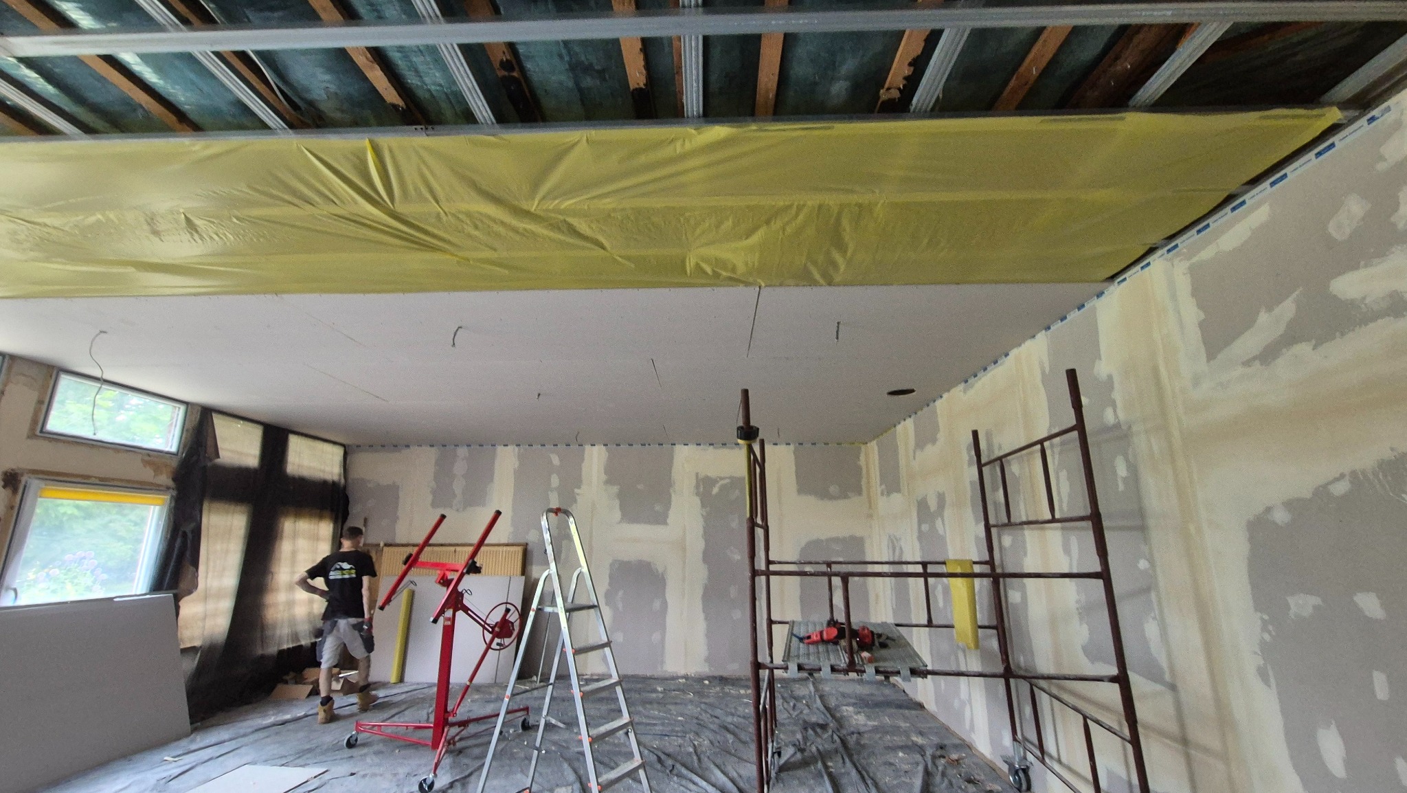 Montaż płyt gipsowo-kartonowych na suficie i ścianach w Legnicy. Widoczny pracownik, rusztowanie i podnośnik do płyt. Etap wykończenia wnętrza.