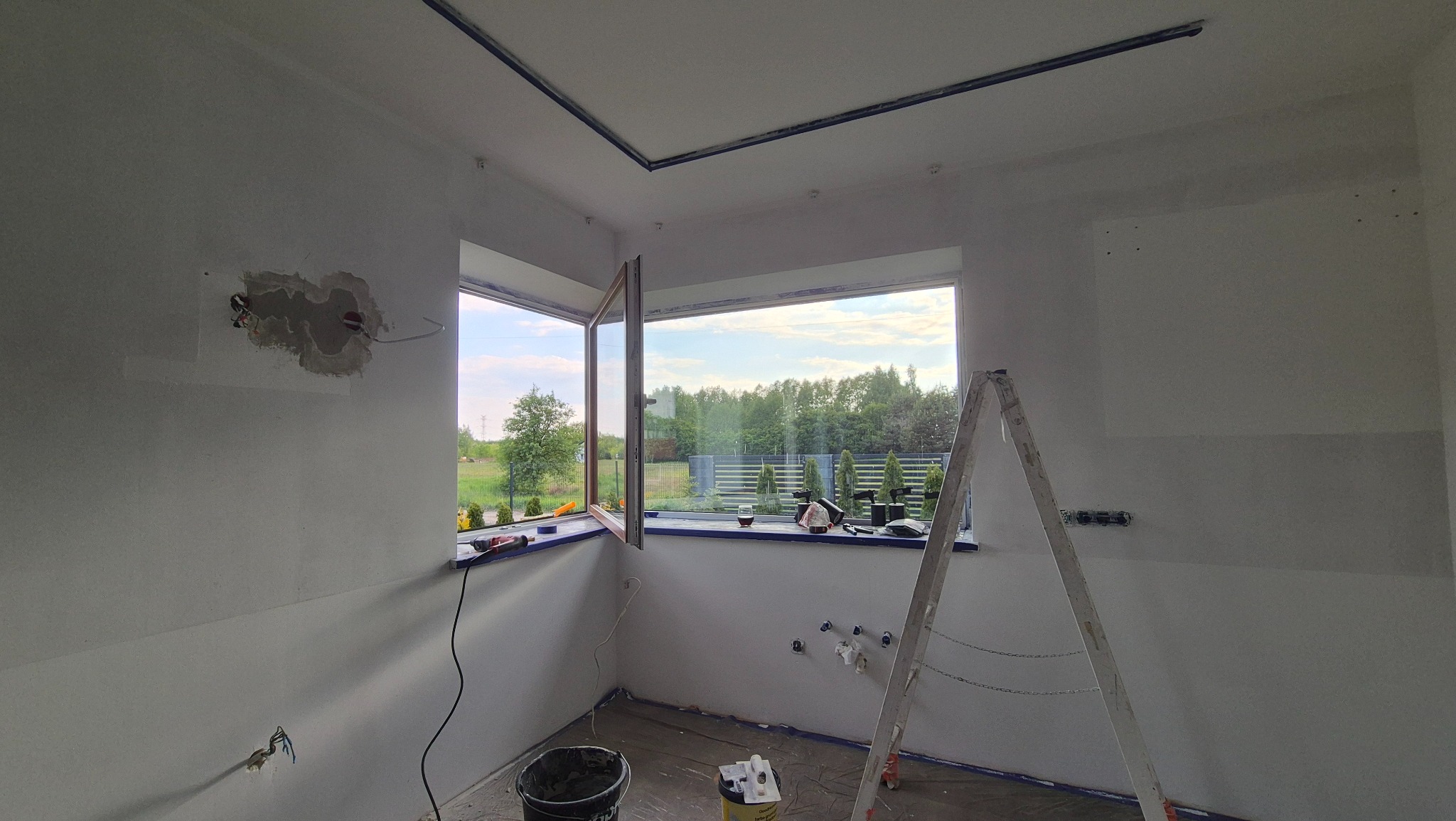 Pokój w trakcie malowania: przygotowane ściany, okna z widokiem na zieleń, drabina malarska, narzędzia. Widoczne niedoskonałości na ścianach przed malowaniem.