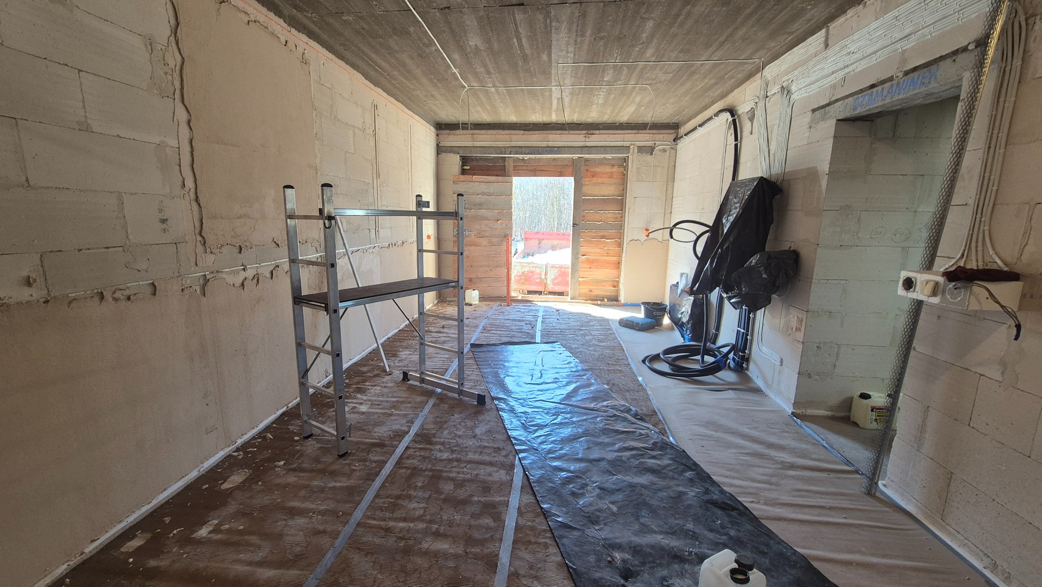 Wnętrze w trakcie prac wykończeniowych: ściany z bloków, częściowo otynkowane, rozłożona folia ochronna, rusztowanie. Widoczne instalacje elektryczne.