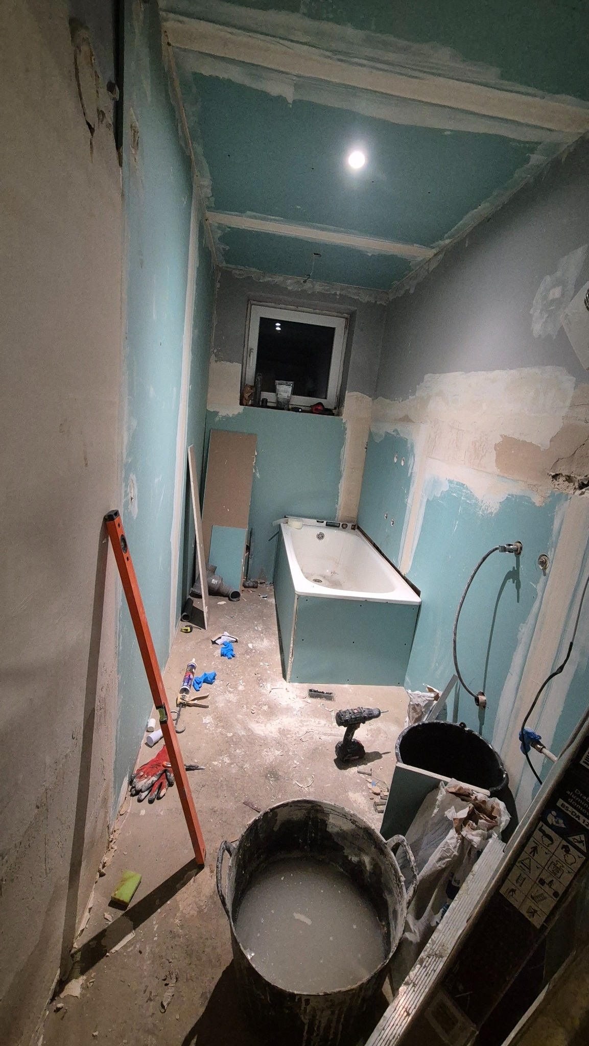 Remont łazienki w trakcie: wanna obudowana płytami, ściany przygotowane do malowania, widoczne narzędzia i materiały budowlane, sufit z płyt gipsowych.