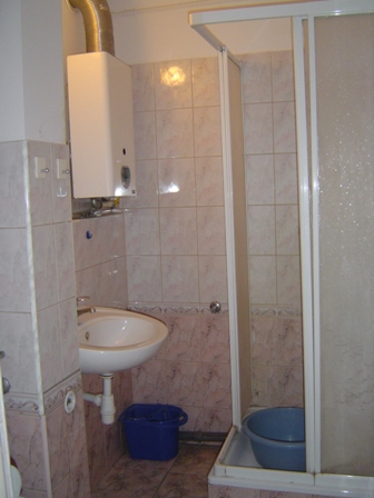 Łazienka z umywalką, piecykiem gazowym i kabiną prysznicową z widocznymi śladami użytkowania, niebieskie wiadro pod umywalką i miska w brodziku.