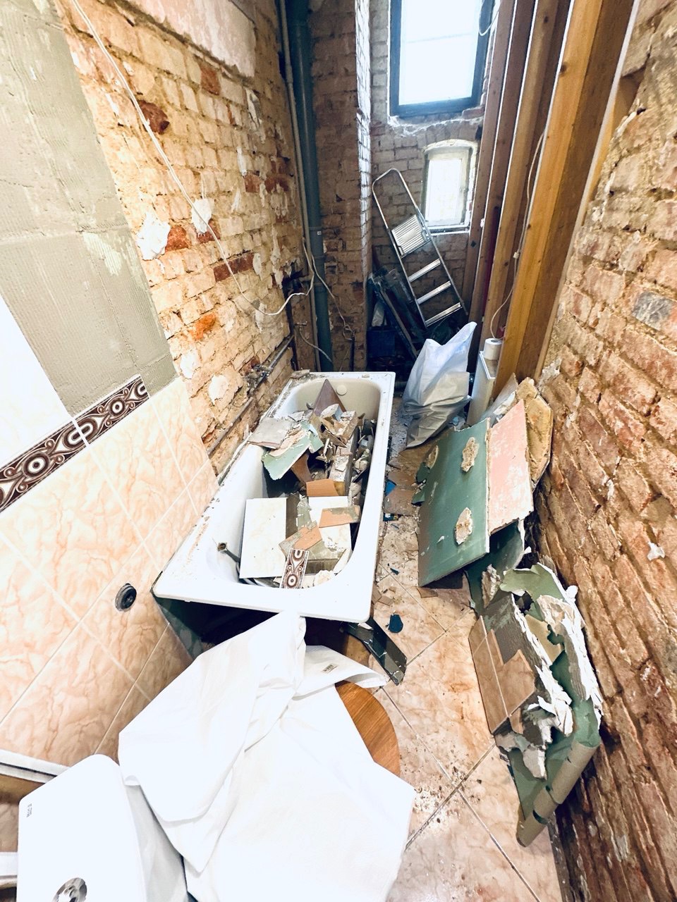 Łazienka w trakcie remontu: odkryta cegła, stara wanna wypełniona gruzem, częściowo położone płytki, drabina oparta o ścianę. Widok na okno.