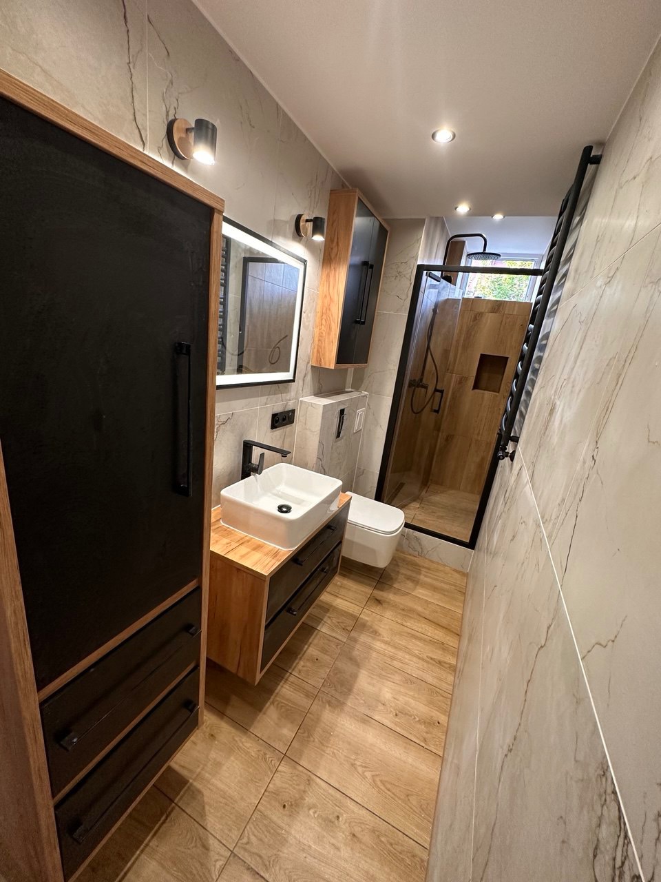 Wykończona, wąska łazienka z kabiną prysznicową typu walk-in, umywalką nablatową na szafce z szufladami, i drewnopodobnymi płytkami na podłodze i w kabinie.