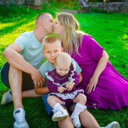 Maciej Aniszewski - Rodzinne zdjęcie plenerowe: rodzice całujący się, synowie siedzą na trawie, dziewczynka trzyma kwiatka. Ujęcie w jasnym, naturalnym świetle, tło z kamiennym murem.