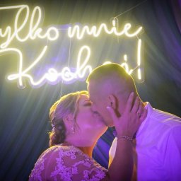 Maciej Aniszewski - Pocałunek pary młodej na tle neonu z napisem 'Tylko mnie kochaj!', fioletowe tło, delikatne światło, widoczne detale sukni ślubnej i biżuterii.