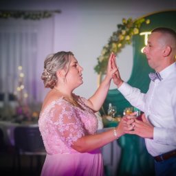 Maciej Aniszewski - Para młoda w trakcie uroczystego przywitania dłońmi na tle dekoracji z kwiatów i neonowego napisu. Panna młoda w różowej sukni z koronką, pan młody w białej koszuli i muszce.