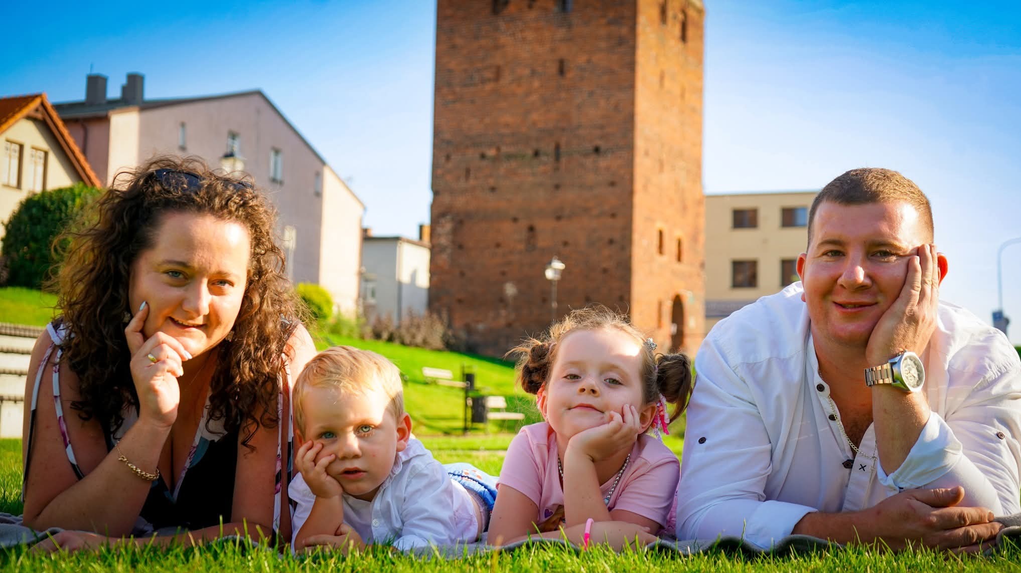 Uśmiechnięta rodzina z dwójką dzieci leży na trawie, na tle ceglanej wieży i budynków. Mama z kręconymi włosami, tata w białej koszuli, dzieci z podpartymi głowami.