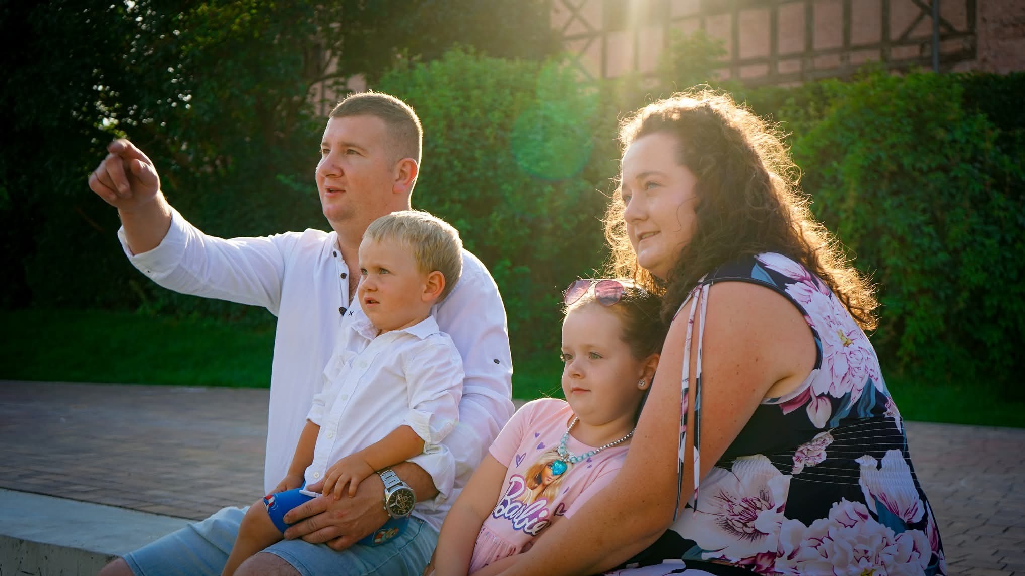 Rodzinne zdjęcie w plenerze: tata wskazuje palcem w górę, mama uśmiecha się, dzieci patrzą w tym samym kierunku. Słońce w tle dodaje ciepła.