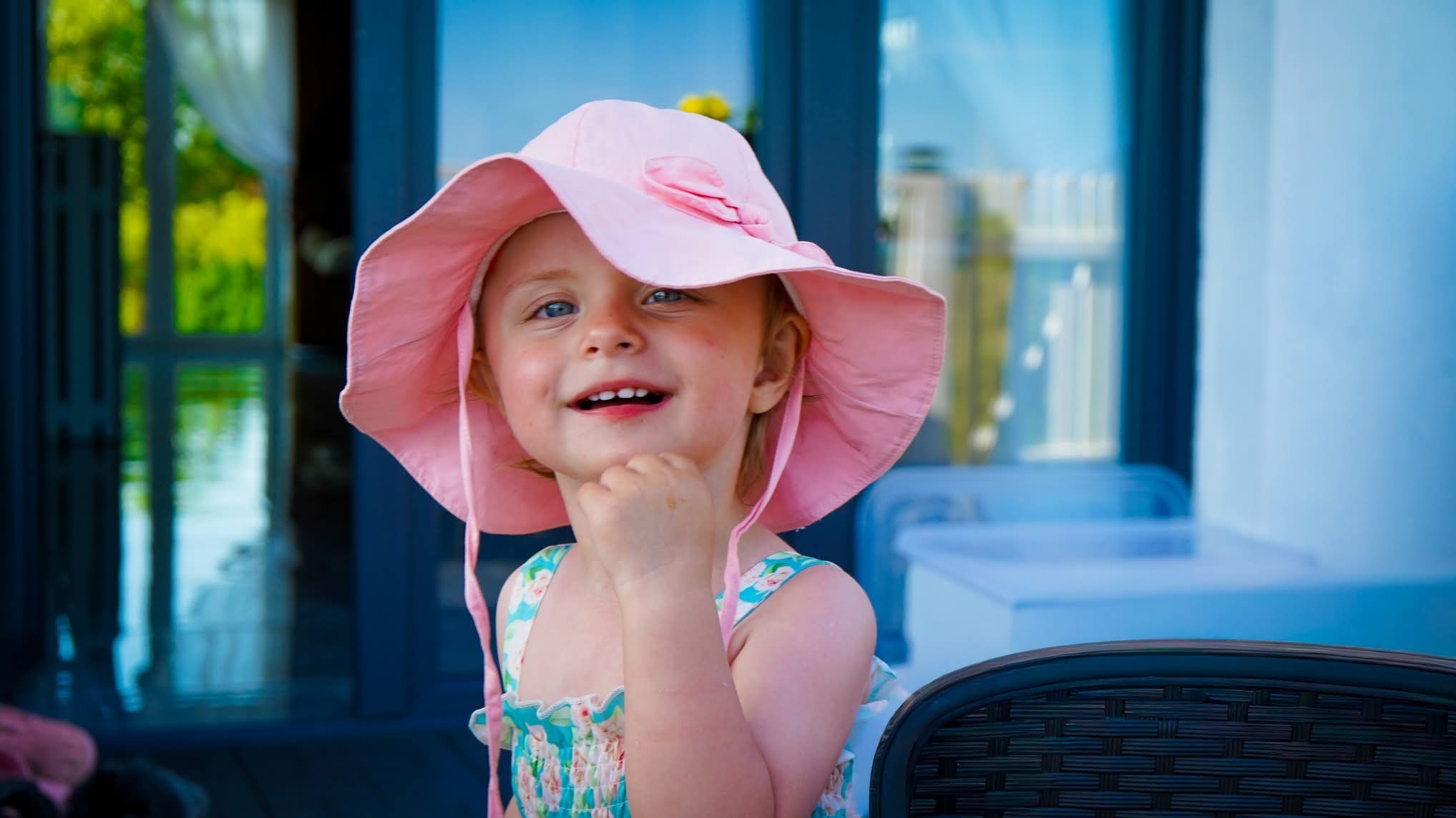 Uśmiechnięta dziewczynka w różowym kapeluszu i kwiecistej sukience, opierająca brodę na dłoni, na tle przeszklonej werandy.