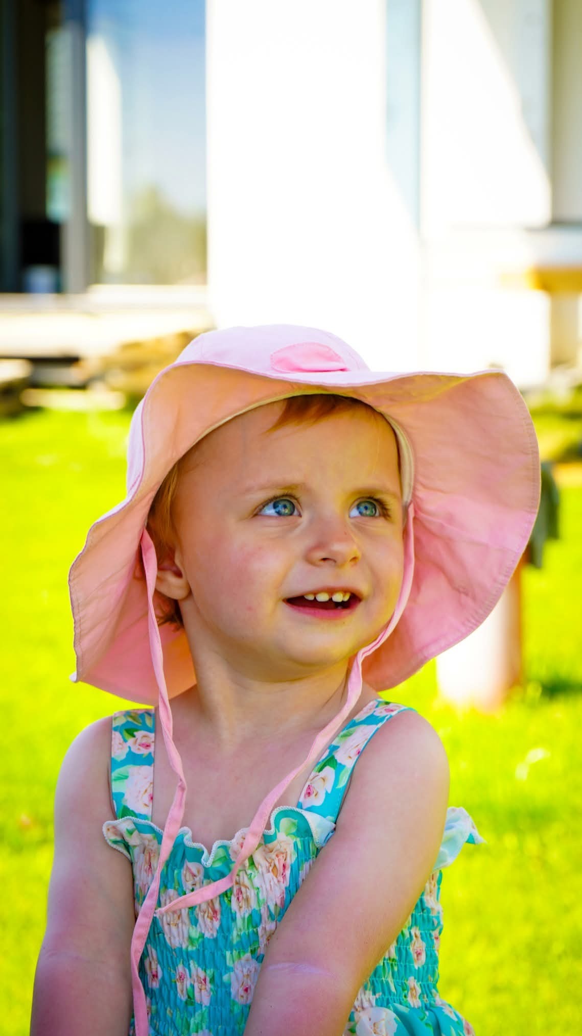 Portret uśmiechniętej dziewczynki w różowym kapeluszu i kwiecistej sukience na tle zielonego trawnika i jasnego budynku.