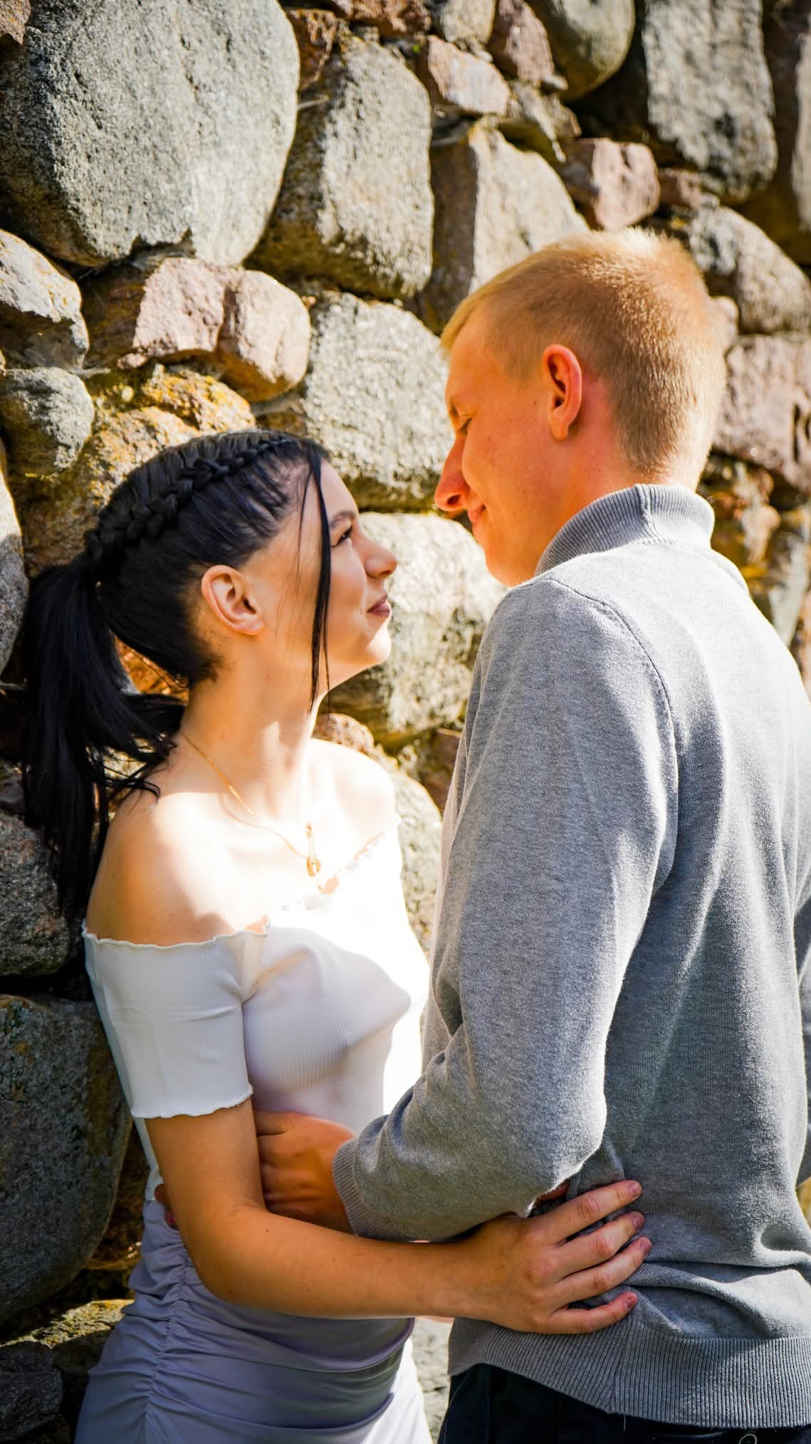 Para zakochanych, brunetka z warkoczami i blondyn w szarym swetrze, przytuleni na tle kamiennej ściany, patrzą sobie w oczy. Ujęcie z bliska, ciepłe światło.