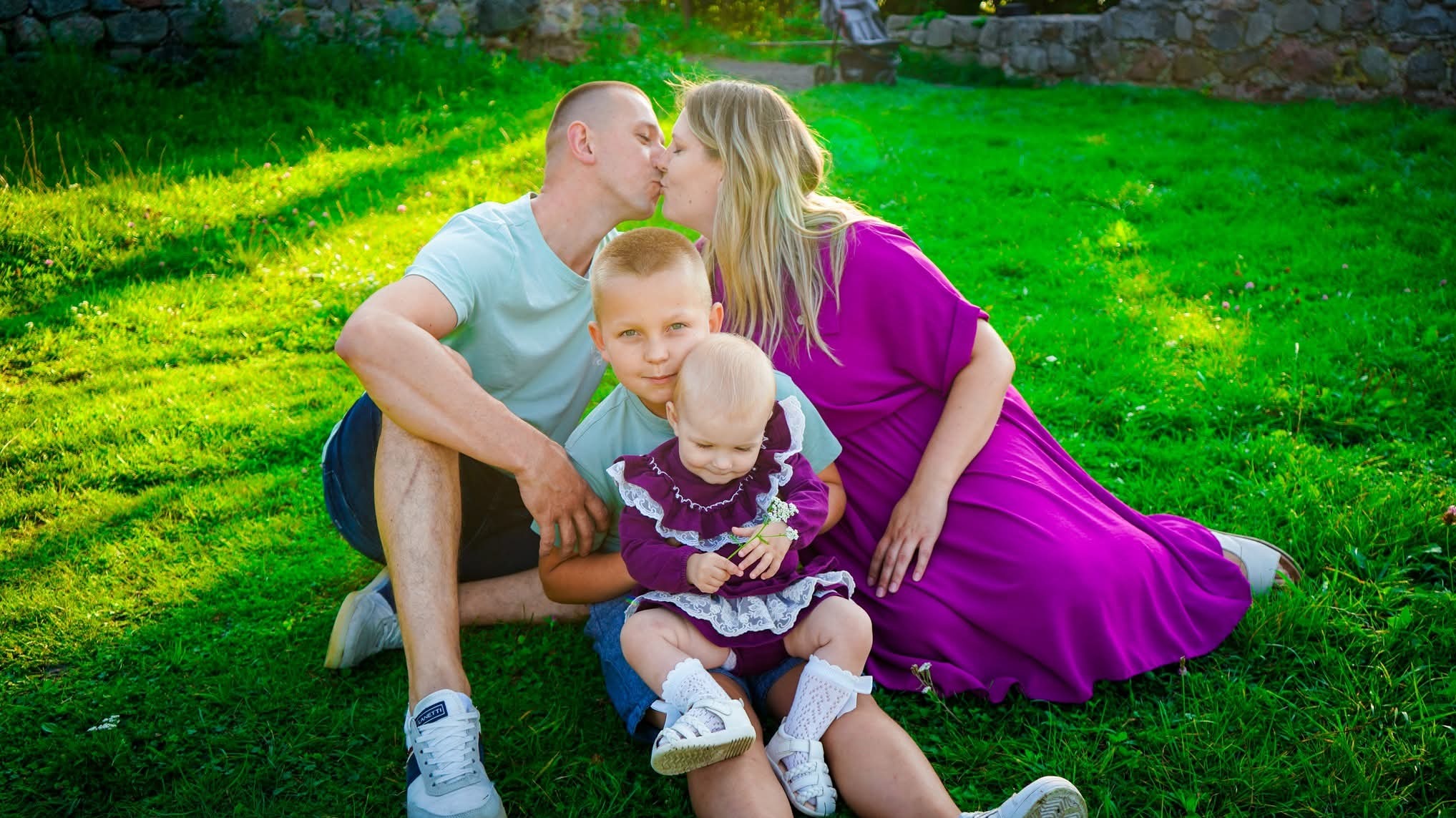 Rodzinne zdjęcie plenerowe: rodzice całujący się, synowie siedzą na trawie, dziewczynka trzyma kwiatka. Ujęcie w jasnym, naturalnym świetle, tło z kamiennym murem.