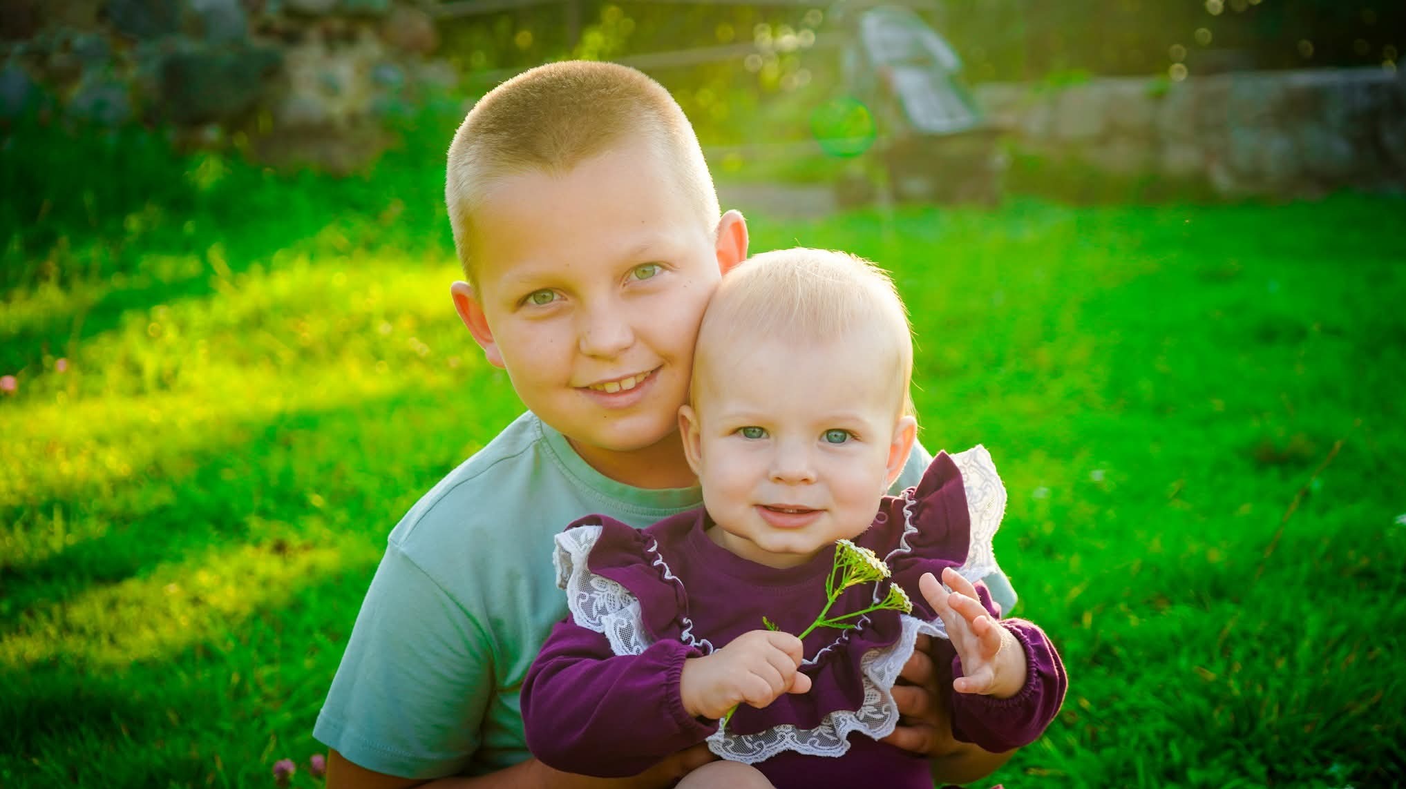 Uśmiechnięty chłopiec obejmuje młodszą siostrę w bordowej sukience z koronką, trzymającą kwiatek na tle zielonej trawy w słoneczny dzień. Portret rodzinny.