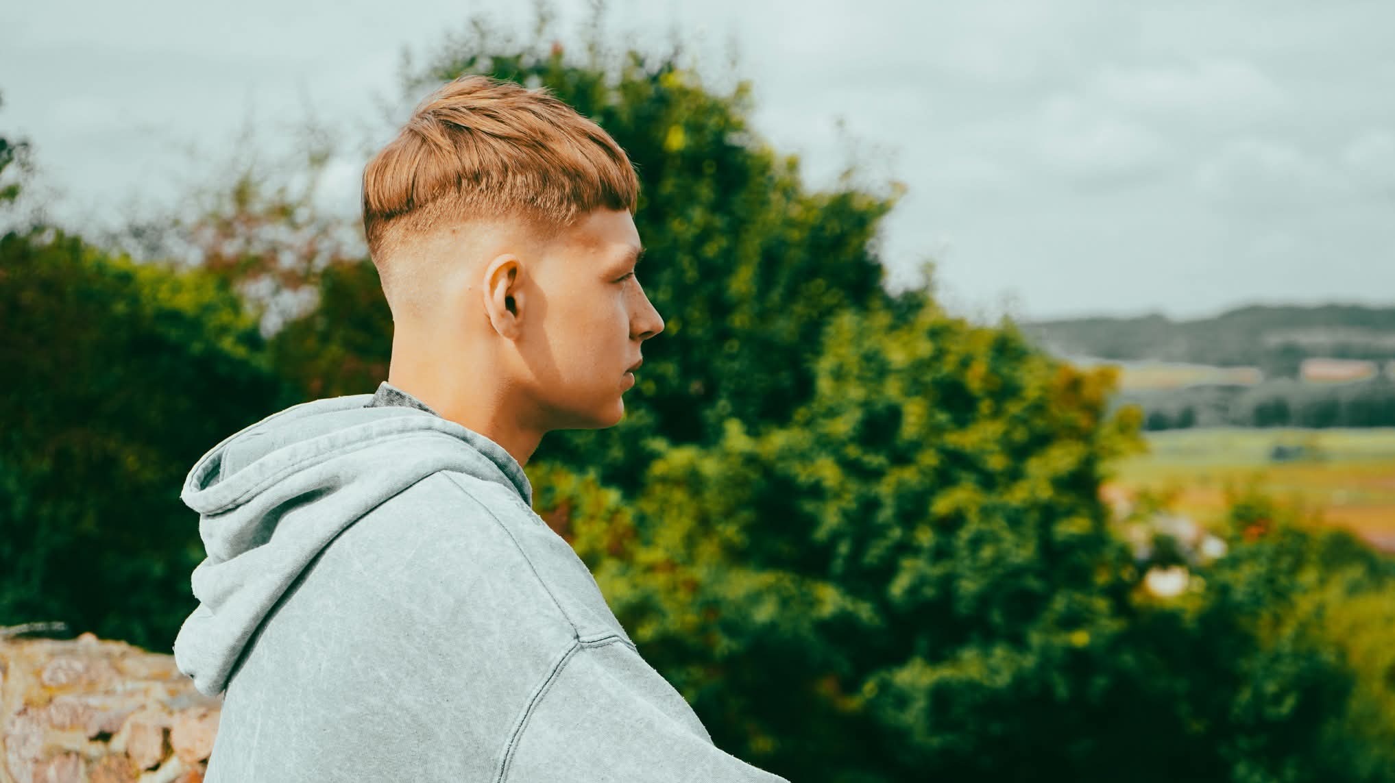 Młody mężczyzna z modną fryzurą side-part w szarej bluzie z kapturem, profil na tle zieleni i rozmytego krajobrazu. Charakterystyczne cięcie włosów.
