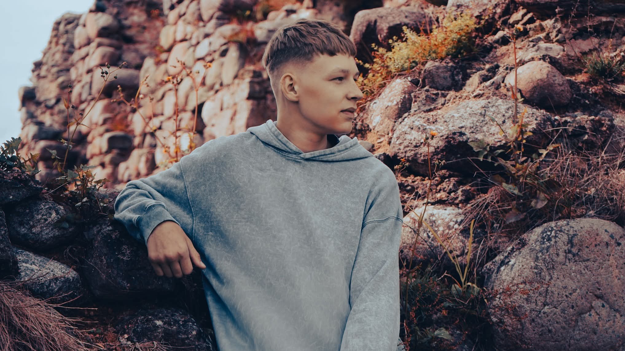 Młody mężczyzna w szarej bluzie z kapturem opiera się o kamienną konstrukcję. Ujęcie z profilu, skupione na twarzy i teksturze kamienia w naturalnym oświetleniu.