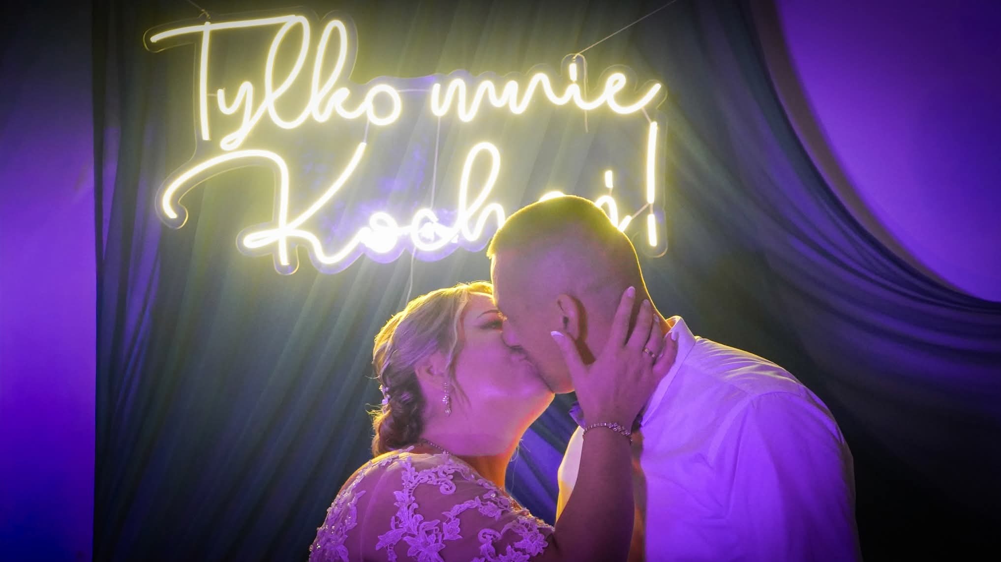 Pocałunek pary młodej na tle neonu z napisem 'Tylko mnie kochaj!', fioletowe tło, delikatne światło, widoczne detale sukni ślubnej i biżuterii.