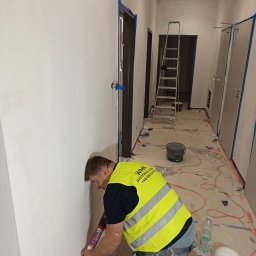 WMK INVESTMENT GROUP SPÓŁKA Z OGRANICZONĄ ODPOWIEDZIALNOŚCIĄ - Pracownik w kamizelce nakłada masę uszczelniającą na ścianę w korytarzu biurowym. Widoczne zabezpieczenia, drabina i przygotowane drzwi do montażu.