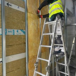WMK INVESTMENT GROUP SPÓŁKA Z OGRANICZONĄ ODPOWIEDZIALNOŚCIĄ - Pracownik w żółtej kamizelce montuje izolację z wełny mineralnej w metalowym stelażu. Widoczne aluminiowe profile i taśmy zabezpieczające. Praca na wysokości z użyciem drabiny.