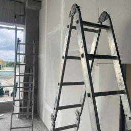 WMK INVESTMENT GROUP SPÓŁKA Z OGRANICZONĄ ODPOWIEDZIALNOŚCIĄ - Dwie drabiny opierają się o ścianę z płyt kartonowo-gipsowych, które są świeżo zamontowane i zaszpachlowane. Widoczne łączenia płyt. Surowy betonowy sufit.