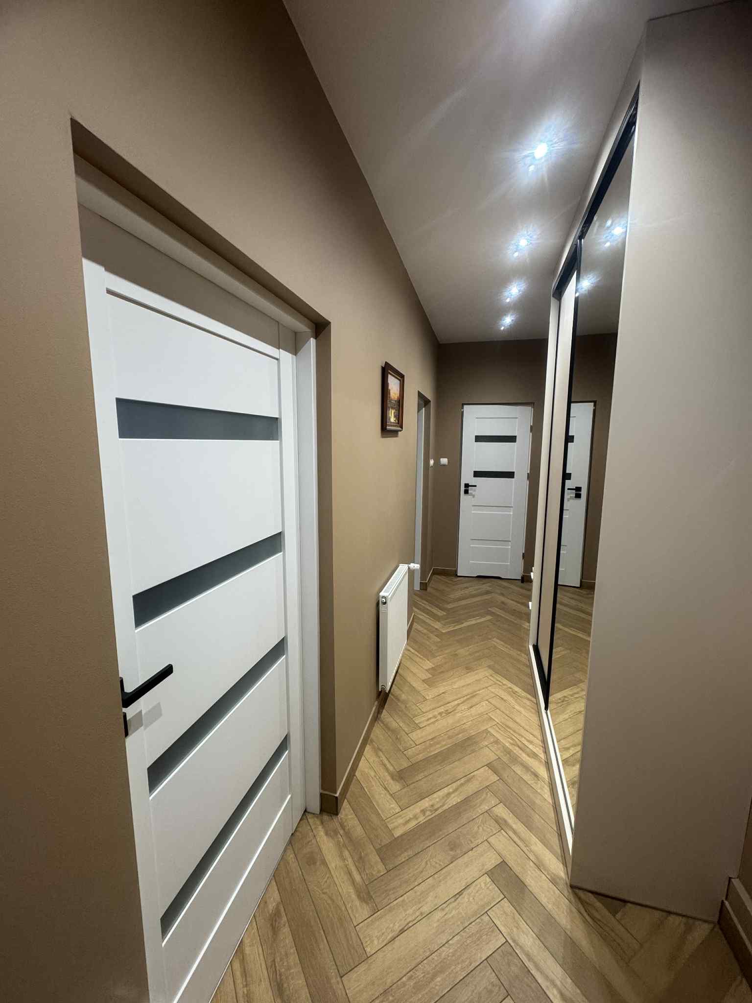 Korytarz po remoncie: białe drzwi z szarymi wstawkami, parkiet w jodełkę, beżowe ściany, szafa z lustrem. Widok na wnętrze mieszkania.