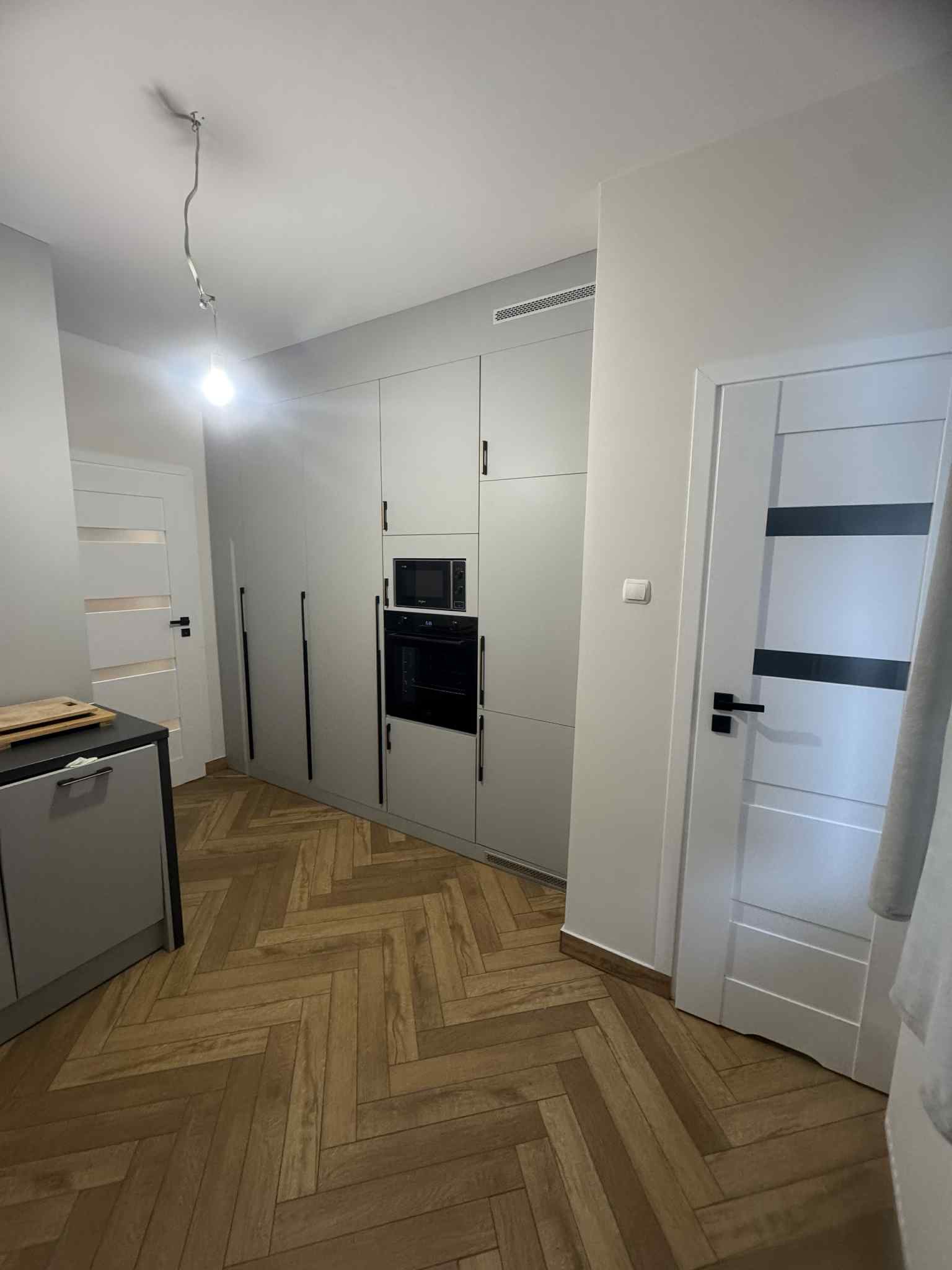 Nowoczesna kuchnia w odcieniach szarości z drewnianą podłogą w jodełkę. Zabudowa kuchenna z piekarnikiem i mikrofalówką. Białe drzwi z czarnymi akcentami.