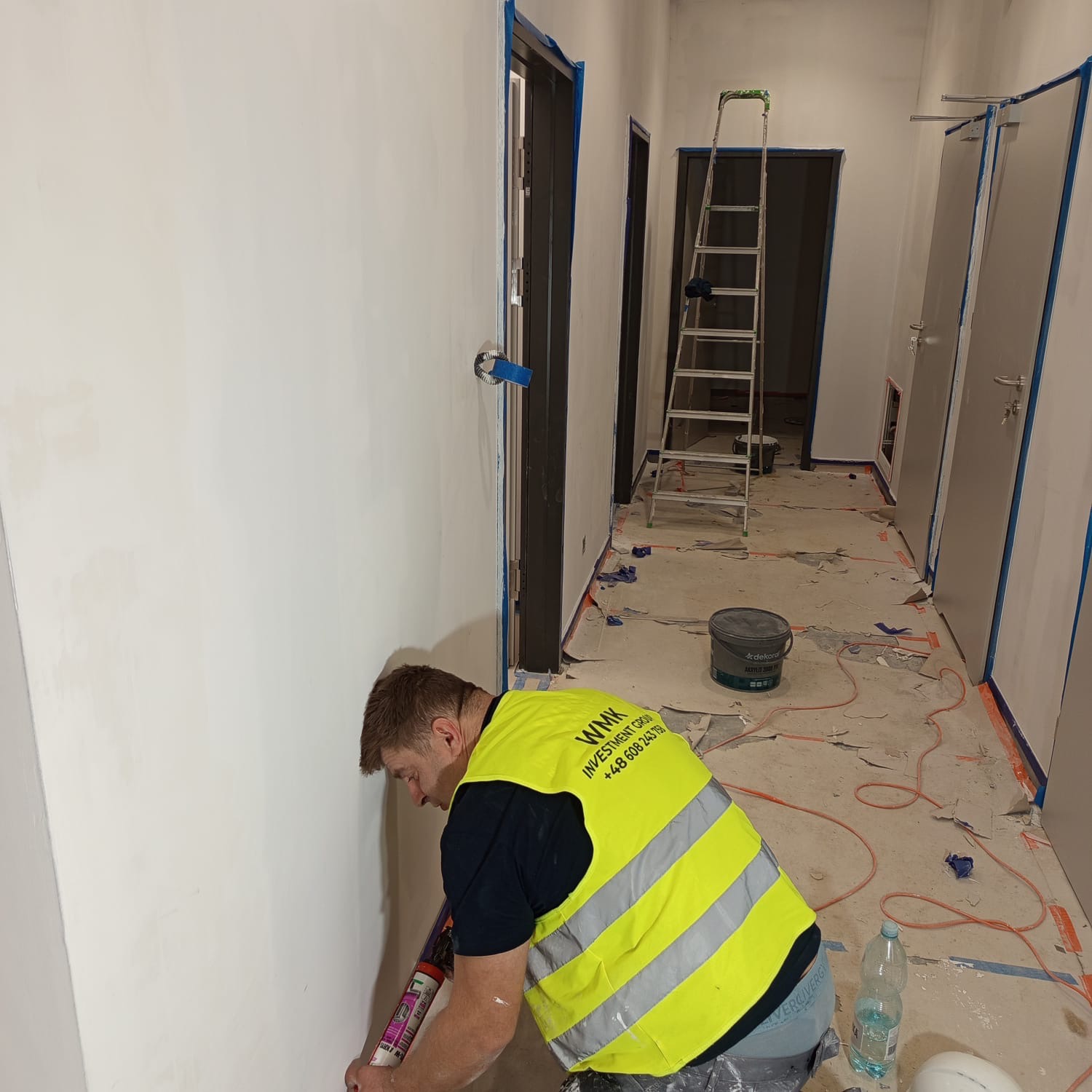 Pracownik w kamizelce nakłada masę uszczelniającą na ścianę w korytarzu biurowym. Widoczne zabezpieczenia, drabina i przygotowane drzwi do montażu.