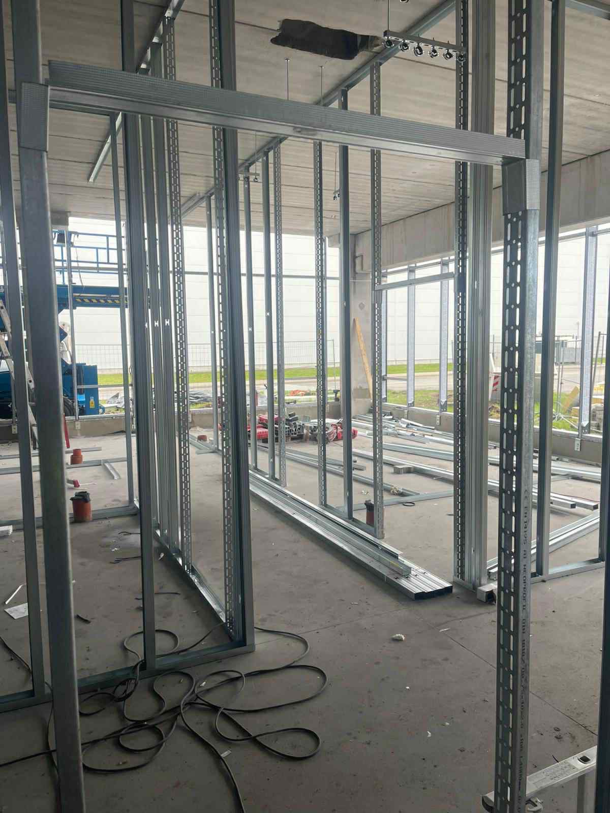 Metalowa konstrukcja ścian działowych z profili, widok z bliska na surowy szkielet przed montażem płyt, narzędzia w tle, surowy beton na podłodze.