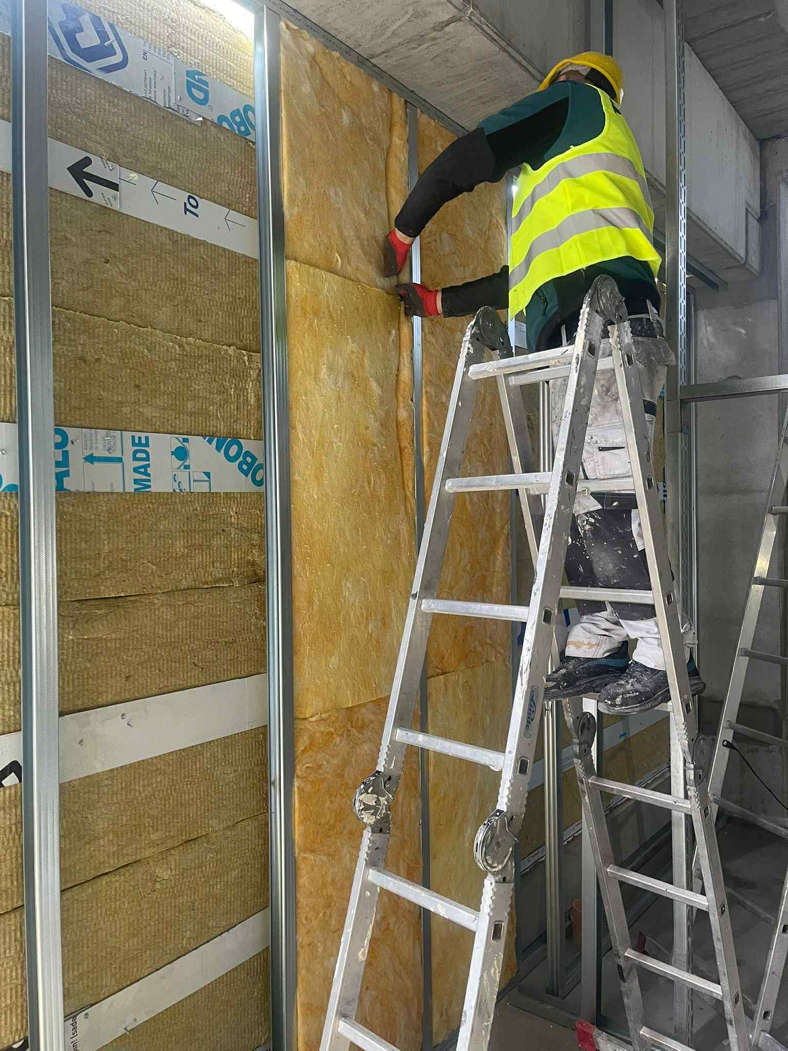 Pracownik w żółtej kamizelce montuje izolację z wełny mineralnej w metalowym stelażu. Widoczne aluminiowe profile i taśmy zabezpieczające. Praca na wysokości z użyciem drabiny.