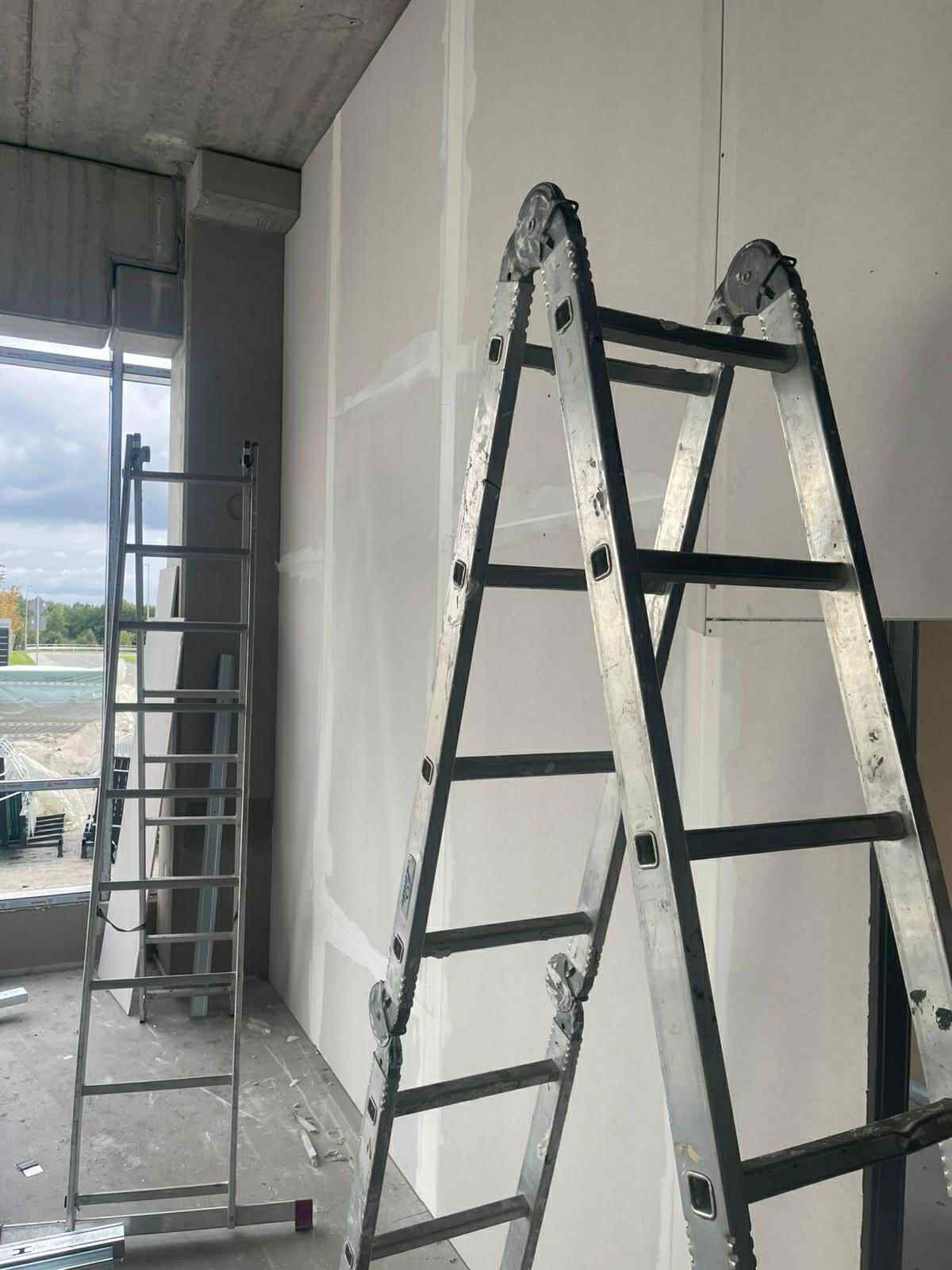 Dwie drabiny opierają się o ścianę z płyt kartonowo-gipsowych, które są świeżo zamontowane i zaszpachlowane. Widoczne łączenia płyt. Surowy betonowy sufit.