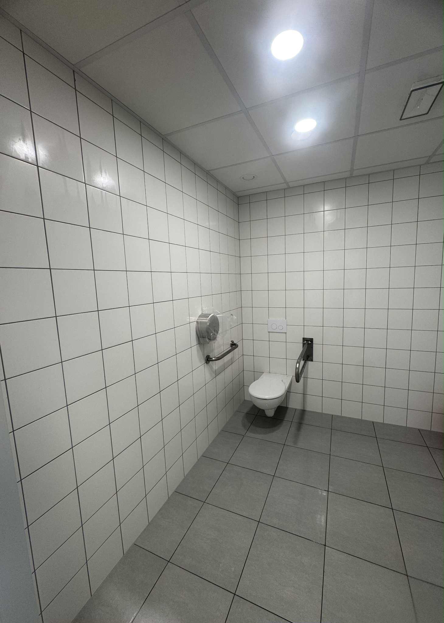 Wykończona toaleta dla niepełnosprawnych z białymi płytkami ściennymi, szarą podłogą i uchwytami. Widoczny podajnik na ręczniki i oświetlenie sufitowe.