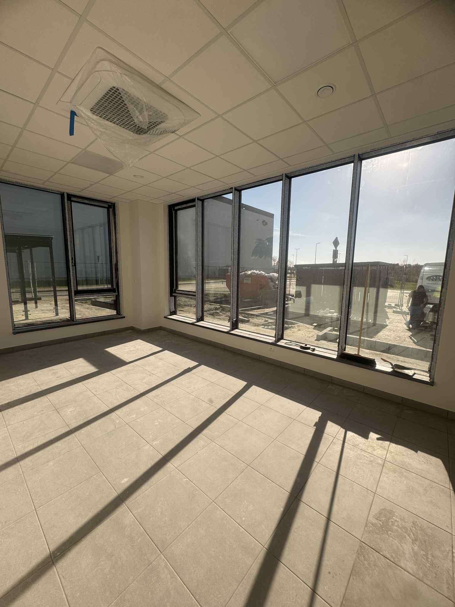 Jasne wnętrze z dużymi oknami i kafelkami na podłodze. Widoczny sufit podwieszany z klimatyzacją. Na zewnątrz budynku widać prace budowlane.