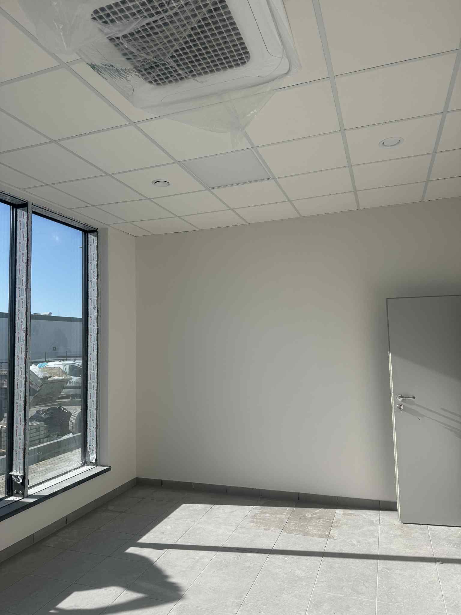 Jasne, puste biuro po remoncie: świeżo pomalowane ściany, podwieszany sufit z kratką wentylacyjną w folii, szare płytki podłogowe, okno z widokiem na zewnątrz i białe drzwi.