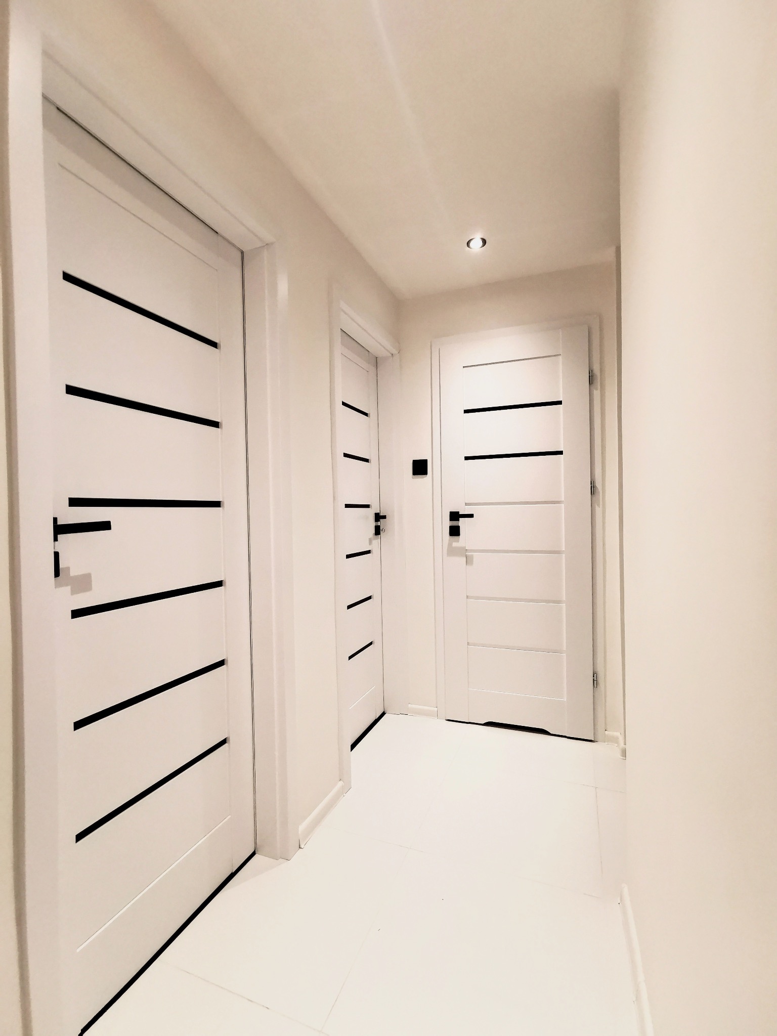 Biały korytarz z trzema nowoczesnymi drzwiami z czarnymi, poziomymi paskami. Minimalistyczny design, jasne oświetlenie, białe ściany i podłoga.