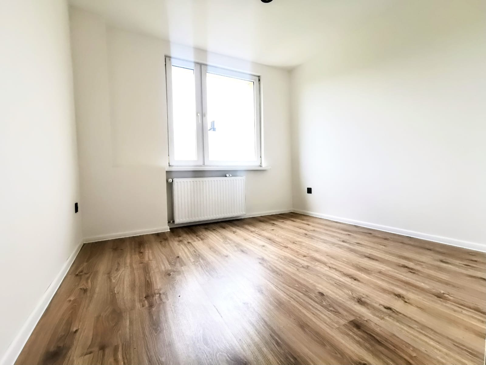 Jasne wnętrze po remoncie: świeżo pomalowane ściany w odcieniach bieli, drewnopodobna podłoga, okno z grzejnikiem. Widok na puste pomieszczenie, gotowe do umeblowania.