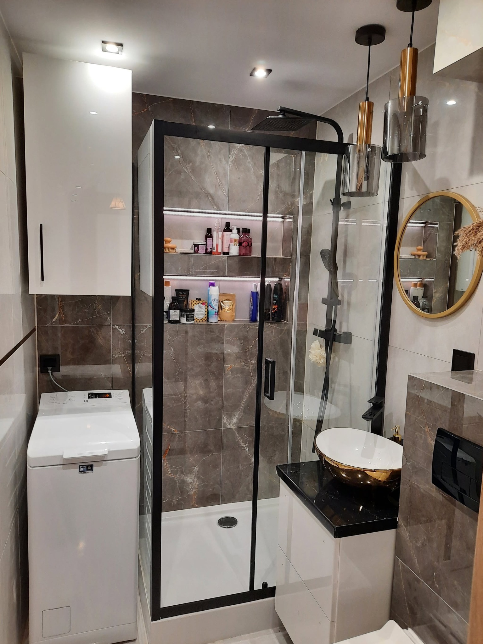 Funkcjonalna mała łazienka po remoncie: kabina prysznicowa typu walk-in z czarną ramą, umywalka nablatowa w kolorze złota, pralka i szafki w nowoczesnym stylu.