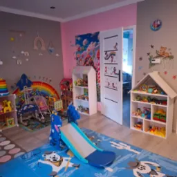 Kolorowy pokój zabaw dla dzieci z zabawkami, regałami w kształcie domków i dywanem z motywami zwierzęcymi. Ściany zdobią naklejki i malunki. W tle otwarte drzwi.