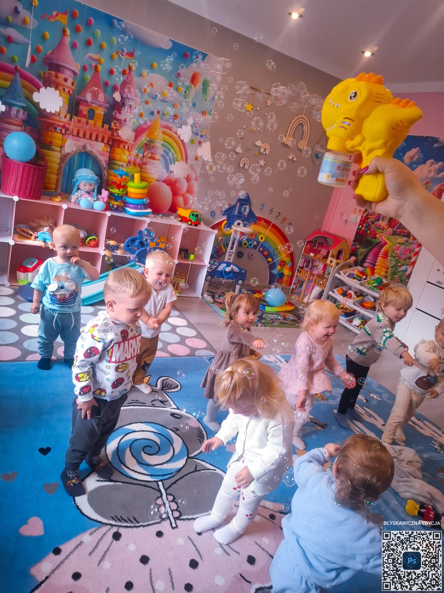 Grupa roześmianych dzieci bawi się w kolorowym pokoju, otoczona bańkami mydlanymi, które puszcza osoba z użyciem zabawki w kształcie żółtego dinozaura.