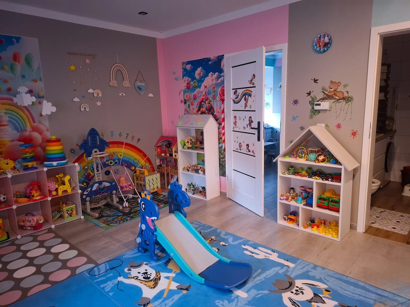 Kolorowy pokój zabaw dla dzieci z zabawkami, regałami w kształcie domków i dywanem z motywami zwierzęcymi. Ściany zdobią naklejki i malunki. W tle otwarte drzwi.