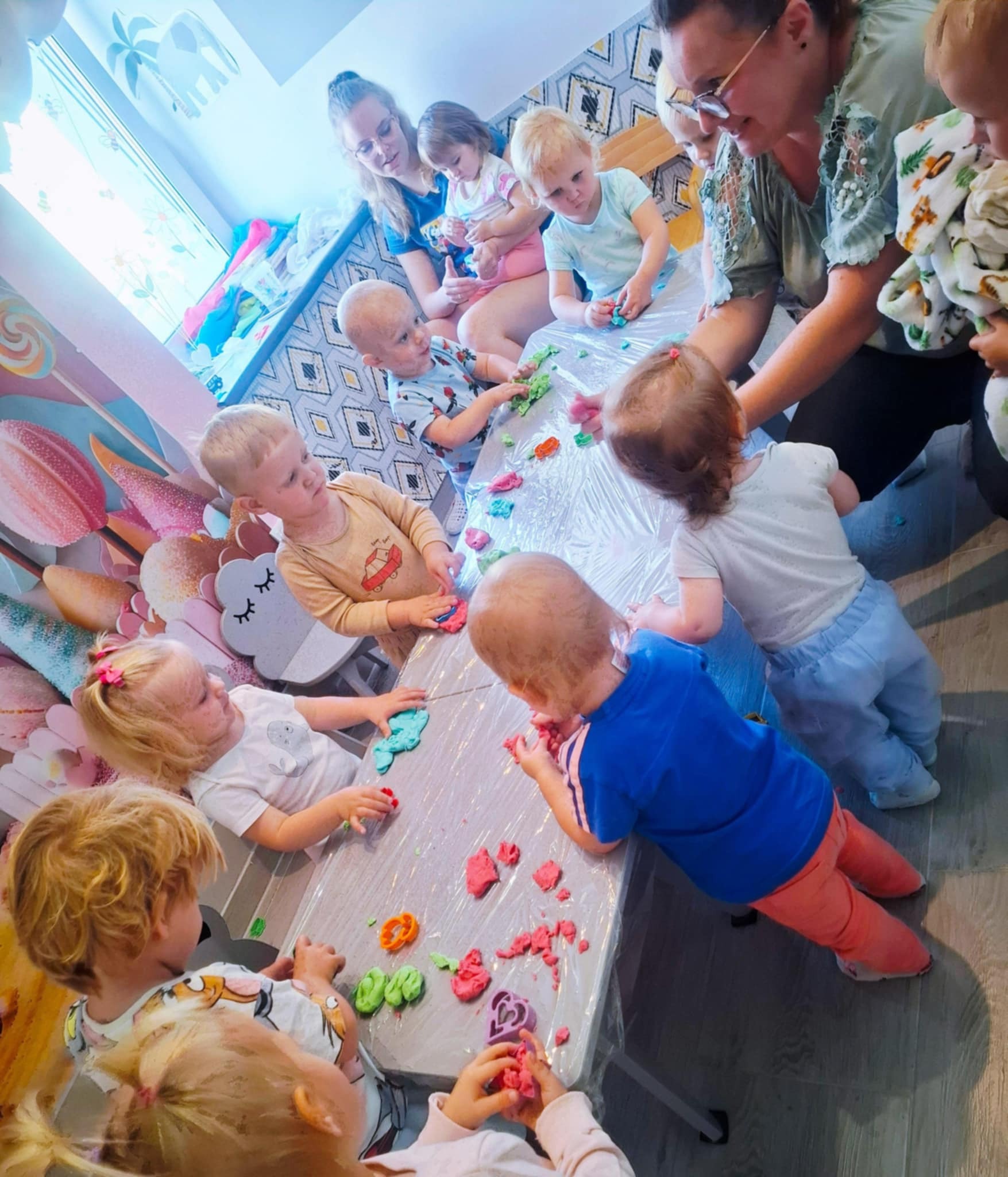 Grupa dzieci w żłobku bawi się kolorową ciastoliną przy stole, nadzorowana przez opiekunki. W tle dekoracje ścian i okien z motywami zwierzęcymi i roślinnymi.