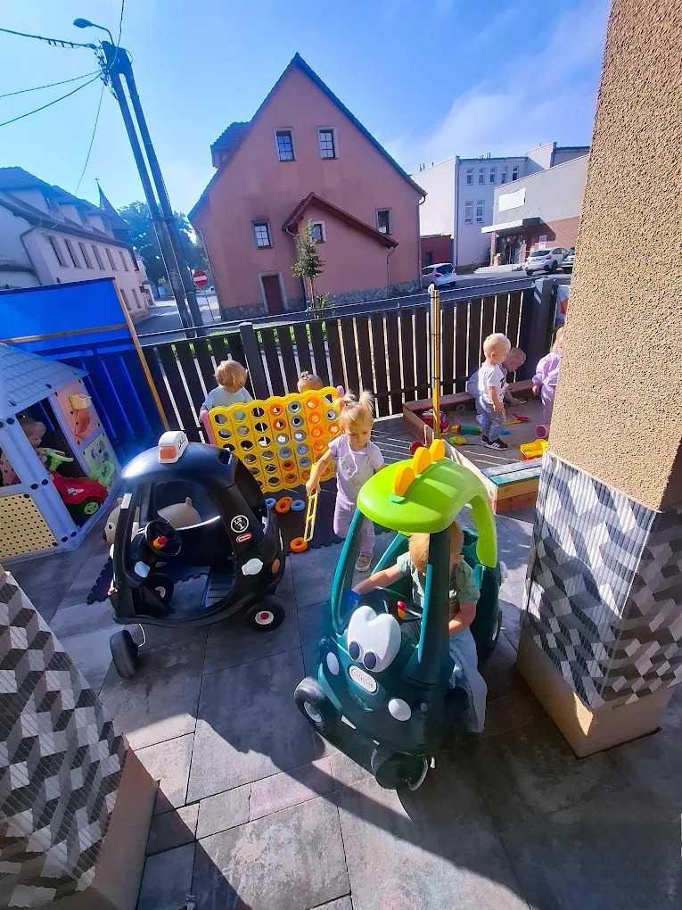 Plac zabaw żłobka z dziećmi bawiącymi się w autkach i grającymi w 'cztery w rzędzie' na świeżym powietrzu, w tle budynki mieszkalne.