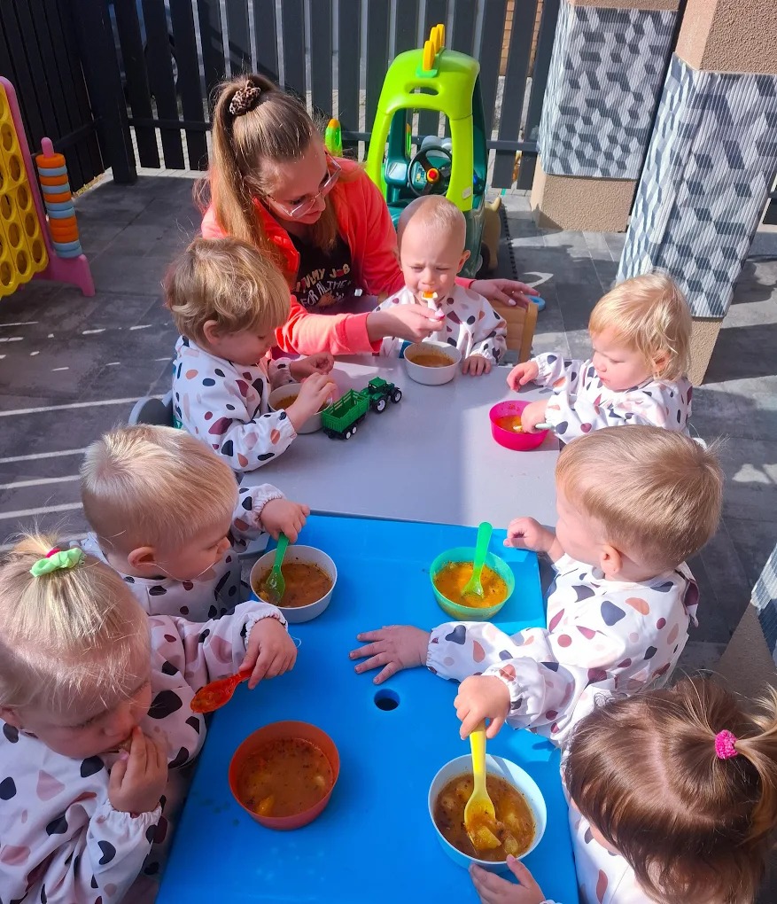 Opiekunka karmi zupą grupę małych dzieci siedzących przy stoliku na zewnątrz. Widoczne kolorowe miseczki i łyżeczki, zabawki i ogrodzenie w tle.