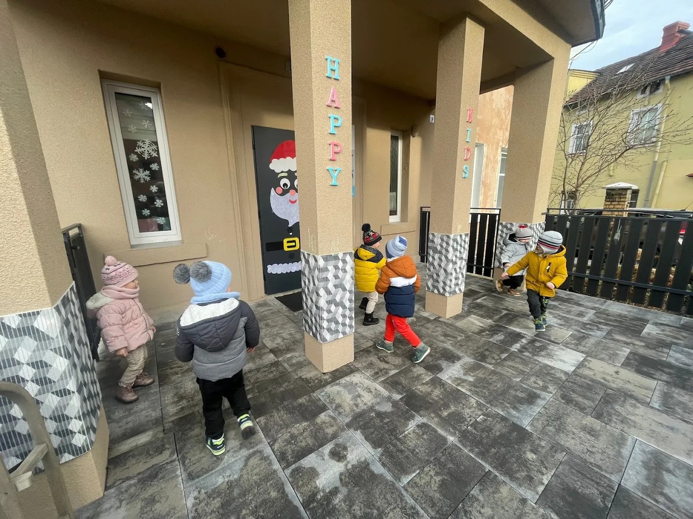 Grupa dzieci w kurtkach wchodzi do budynku z napisem 'HAPPY KIDS' i dekoracją świąteczną na drzwiach. Widoczny chodnik z szarej kostki i filary z wzorem.