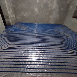 Krisbaum - Niebieskie rury ogrzewania podłogowego ułożone na siatce w surowym pomieszczeniu, gotowe do zalania betonem. Widok z góry, instalacja w trakcie realizacji.