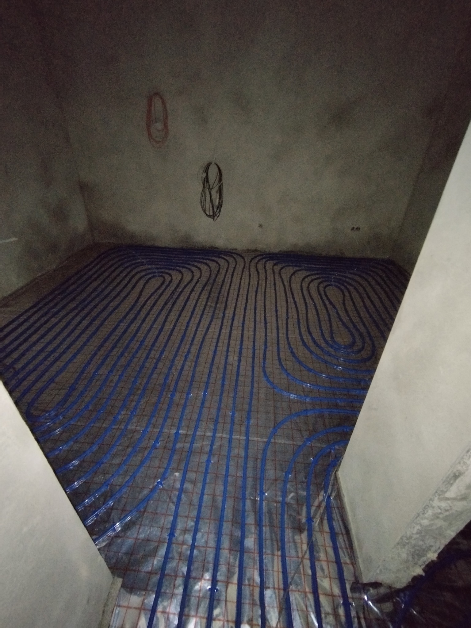 Niebieskie rury ogrzewania podłogowego ułożone na siatce w surowym pomieszczeniu z widocznymi przewodami elektrycznymi na ścianie.