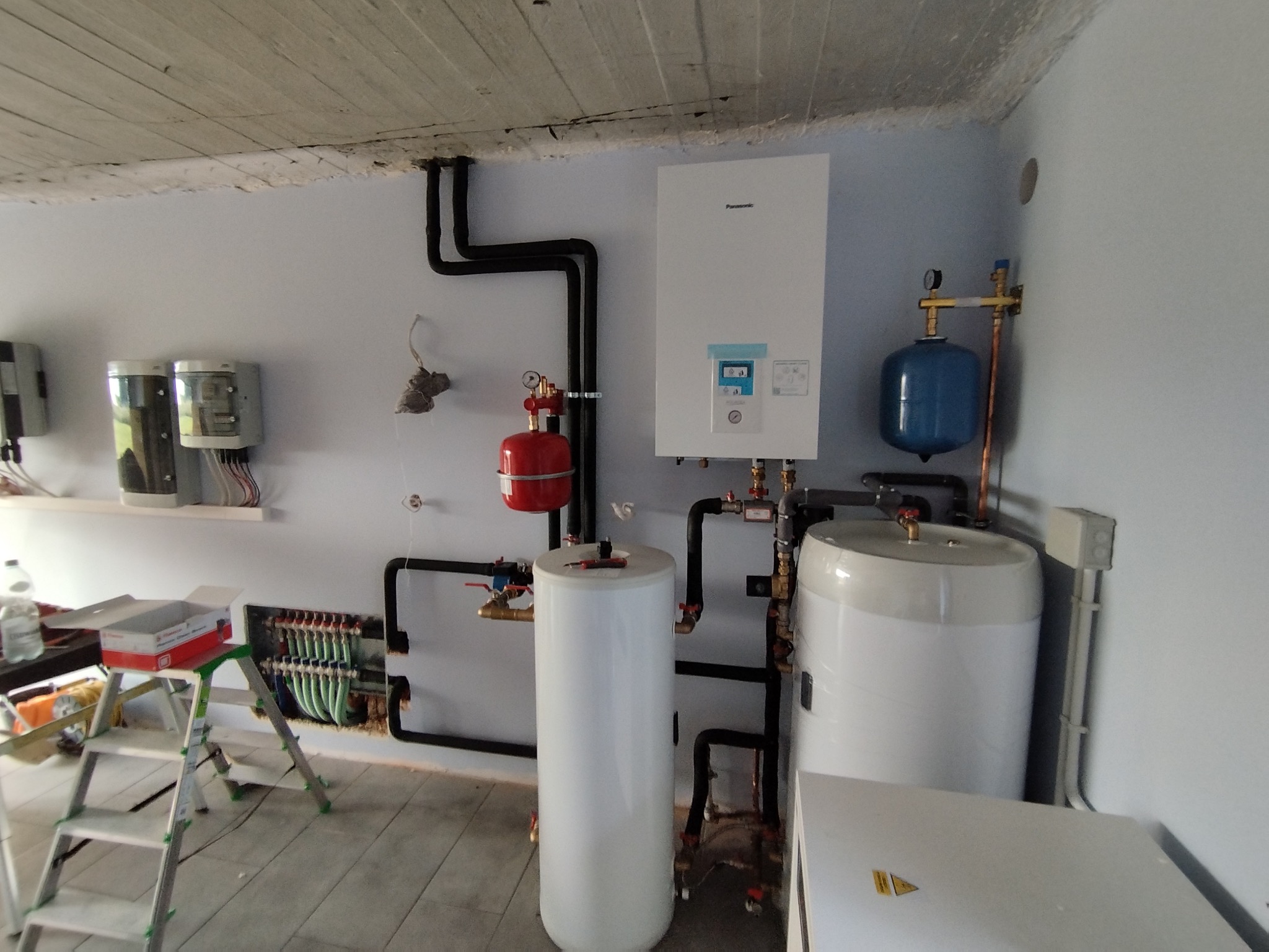 Złożona instalacja grzewcza z kotłem Panasonic, zbiornikami i rurami w piwnicy. Widoczny rozdzielacz ogrzewania podłogowego i drabina monterska.