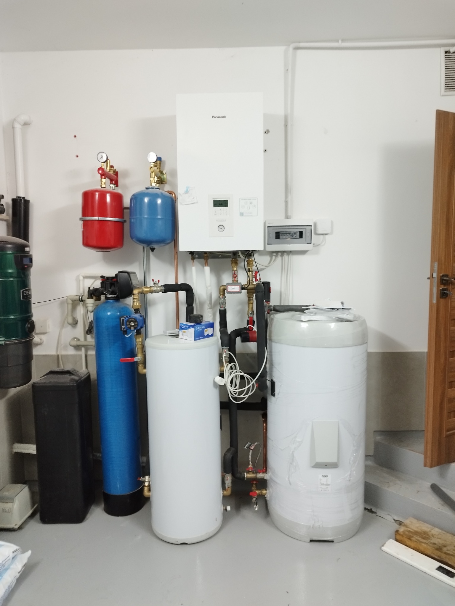 Zestaw urządzeń w kotłowni: biały piec Panasonic, zbiorniki (czerwony, niebieski, białe), filtry. Widoczne rury, zawory i przewody. Szara podłoga, białe ściany.